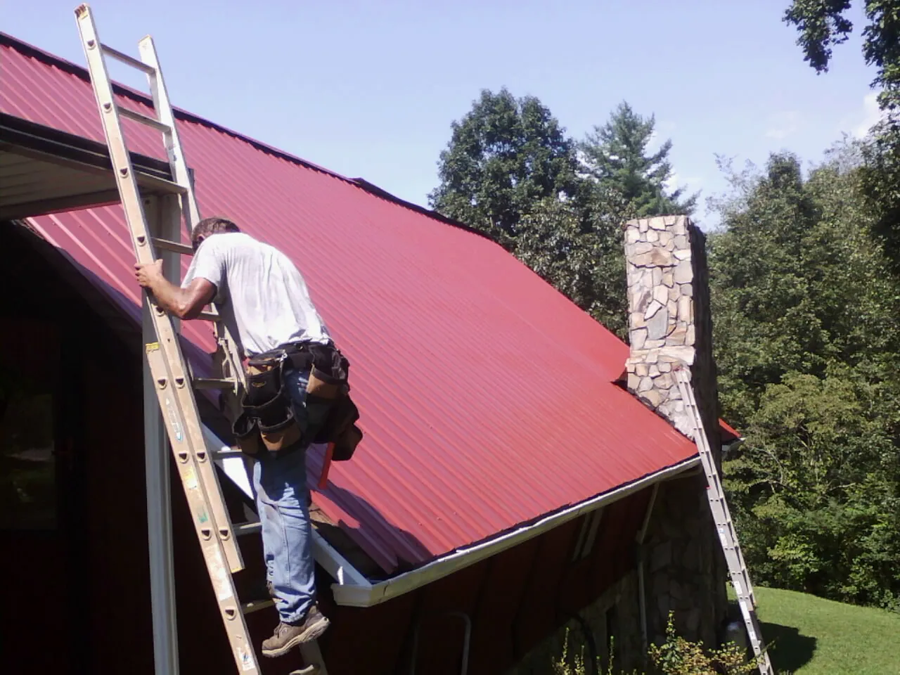 Roofer on a ladder beside a red metal roof and stone chimney — Soffit & Fascia Repair by Roofing Brothers in Clinton Township